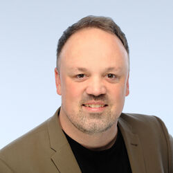 Porträt eines Mannes mit dunklen Haaren. Er trägt ein schwarzes Shirt und ein braunes Sakko.