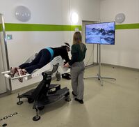 Eine Person liegt auf einer Art Liege, trägt eine VR Brille und schaut in Richtung eines Bildschirms. Daneben steht eine Person die auch in Richtung des Bildschirms blickt.