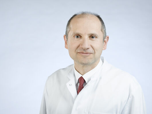 Porträtfoto eines Mannes mit kurzen Haaren und einer hohen Stirn. Unter seinem weißen Arztkittel trägt er ein weißes Hemd und eine rote Krawatte. Er lächelt freundlich in die Kamera. 