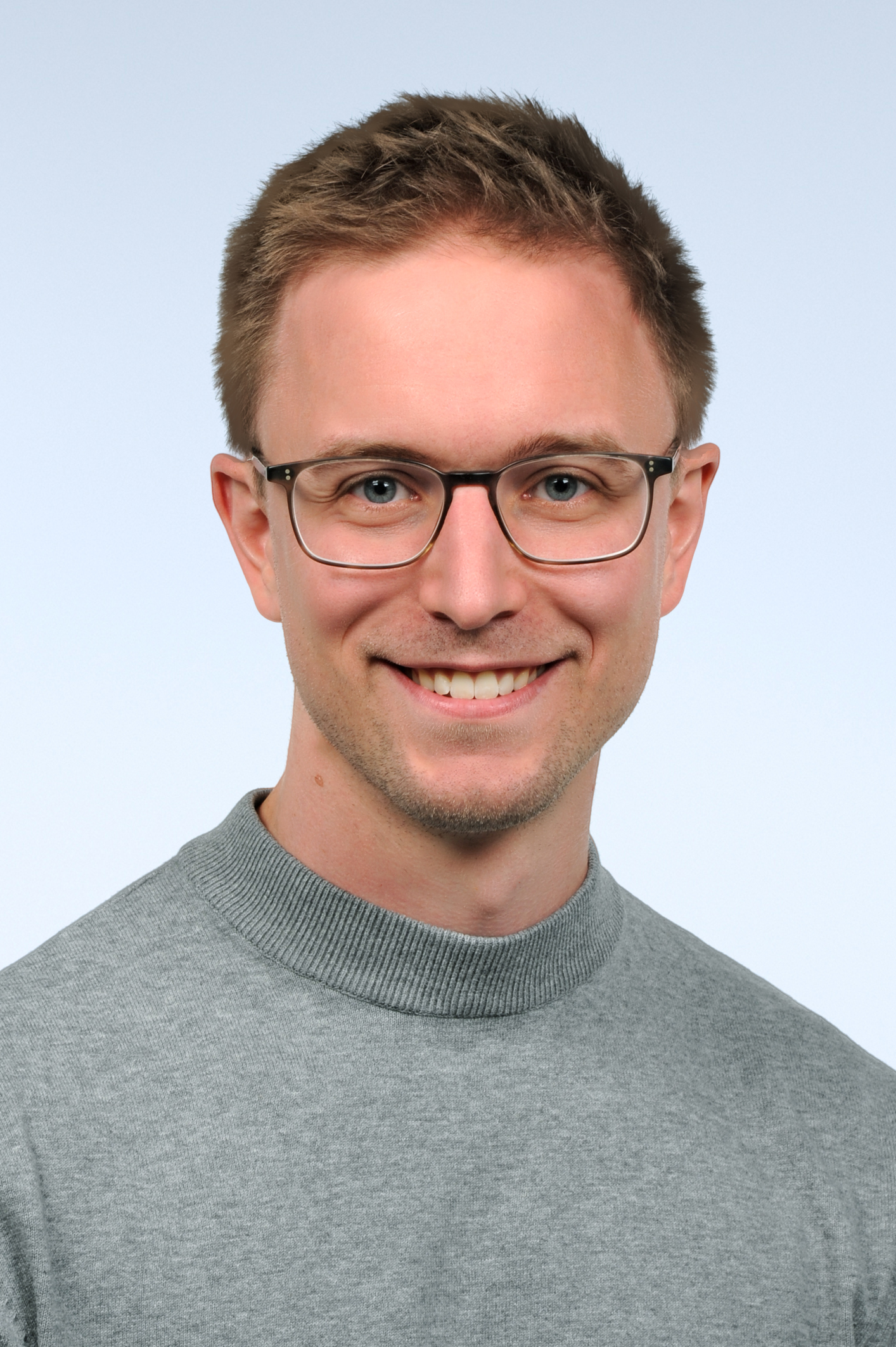 Portraitfoto von Mann mit kurzen dunkelblonden Haaren und Brille. Er trägt einen grauen Pullover und lacht in die Kamera.