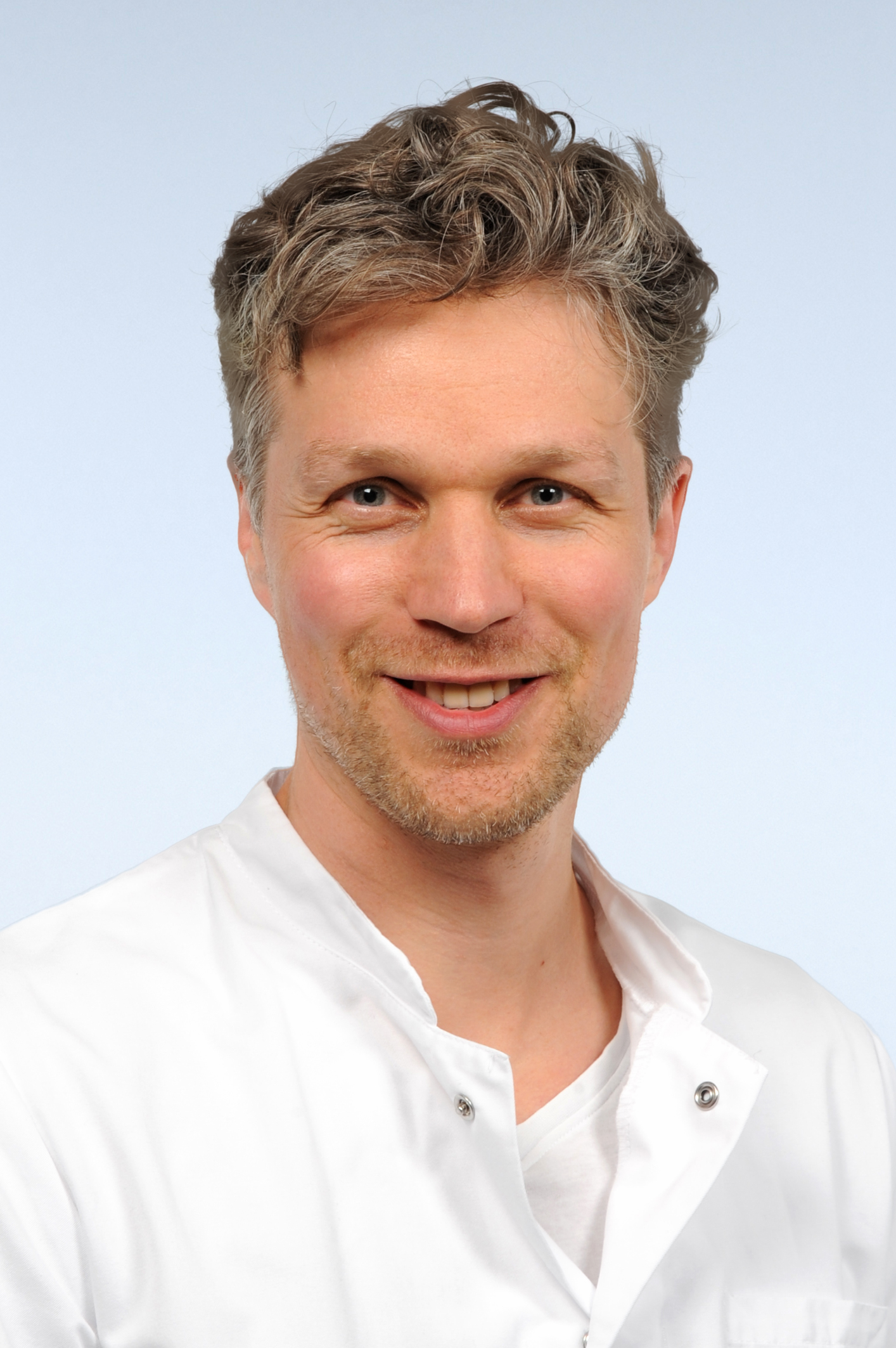 Portraitfoto von Mann mit kurzen, grauen, lockigen Haaren und Dreitagebart. Er lacht in die Kamera und trägt einen weißen Arztkittel.