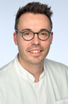 Portraitfoto eines Manns mit kurzen braunen Haaren und runder Brille, der freundlich in die Kamera lächelt.