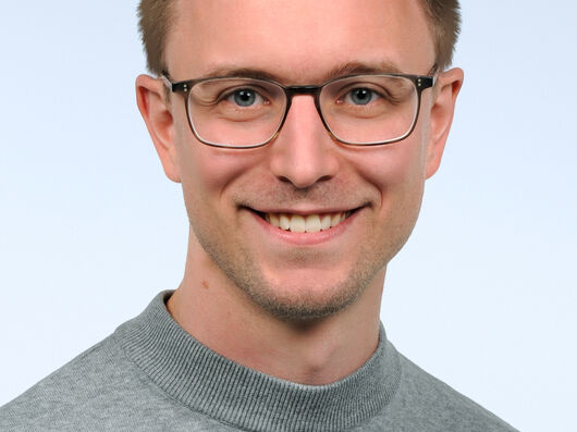 Dr. Dennis Eschweiler Portraitfoto von Mann mit kurzen dunkelblonden Haaren und Brille. Er trägt einen grauen Pullover und lacht in die Kamera.