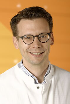Portraitfoto von einem Mann mit braunem Kurzhaarschnitt und runder Brille, er trägt einen Arztkittel und lacht in die Kamera.