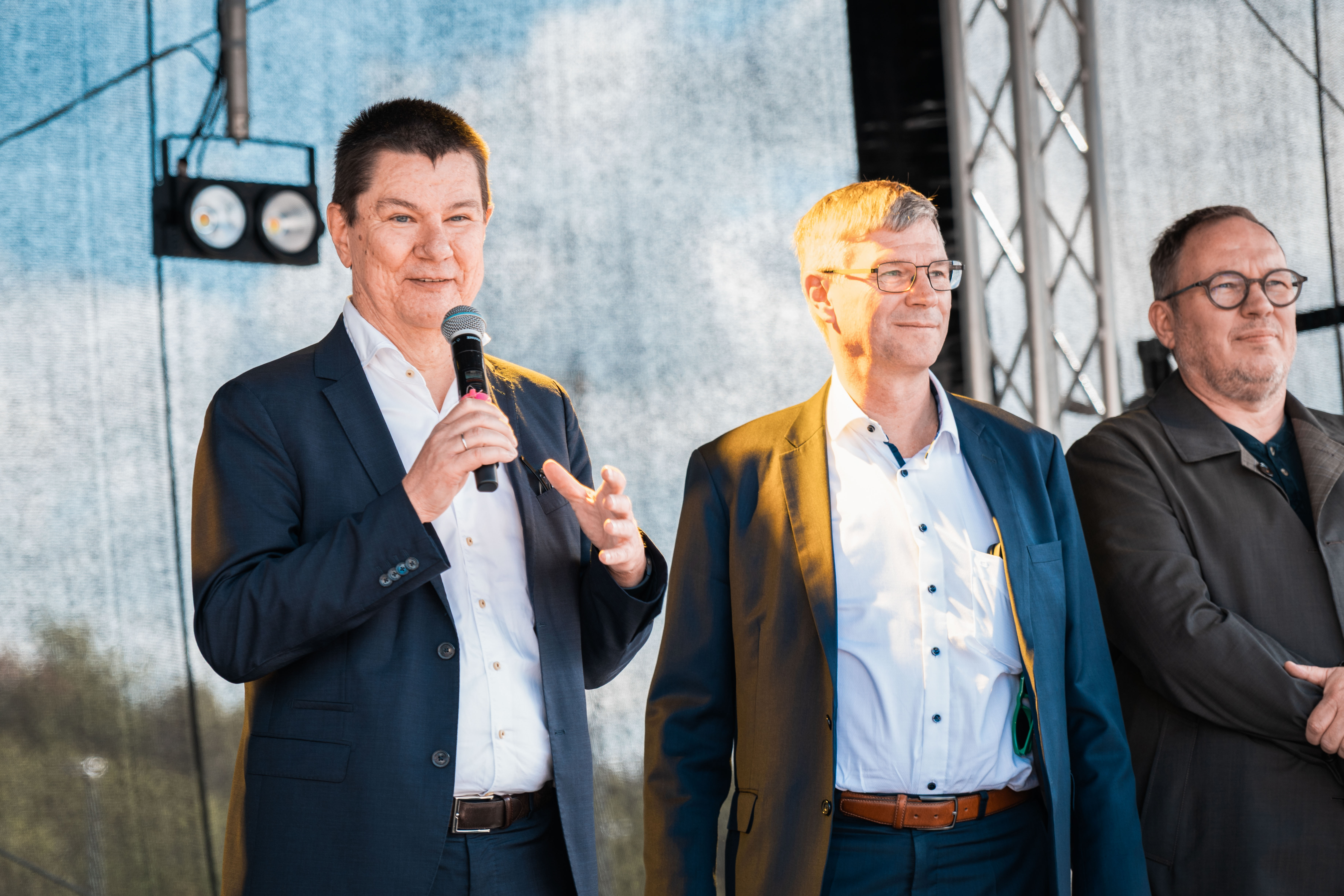 Drei Männer in Business Kleidung stehen auf einer Bühne, einer von ihnen spricht in ein Mikrofon.