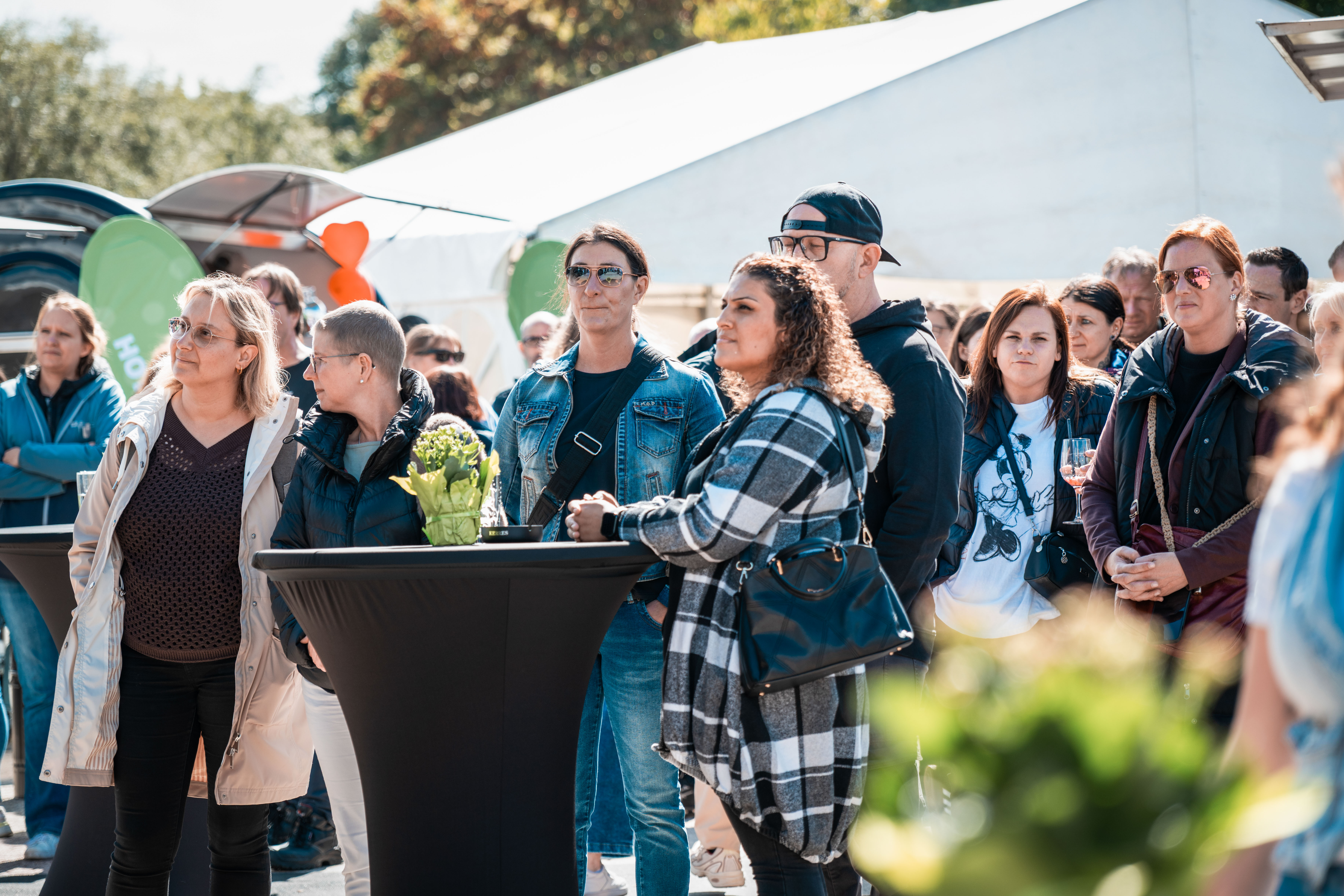 Viele Menschen stehen vor einer Bühne an mehreren Stehtischen und hören dem Programm auf der Bühne zu.