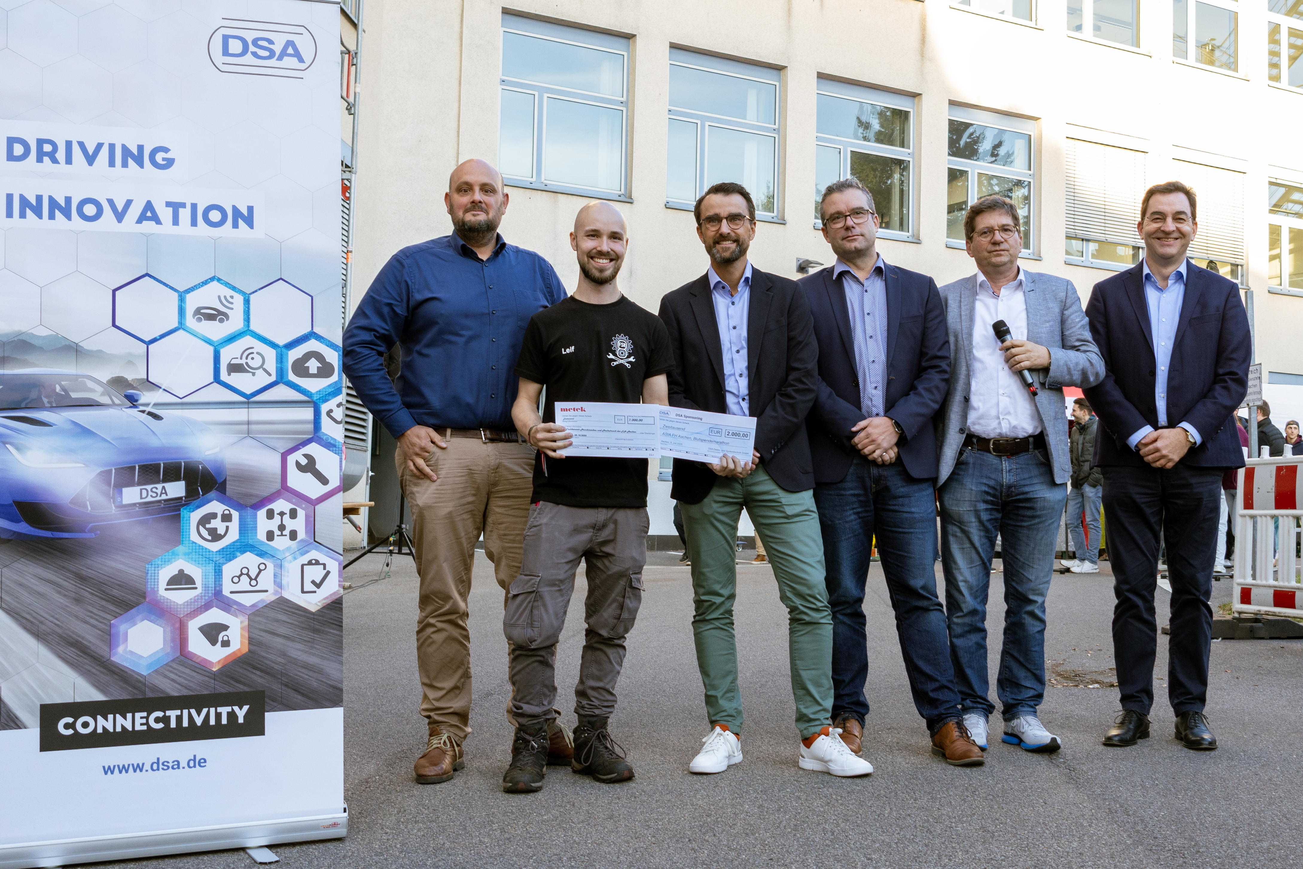 Gruppenaufnahme von sechs Männern, die freundlich in die Kamera lächeln. Zwei Männer halten einen Scheck.