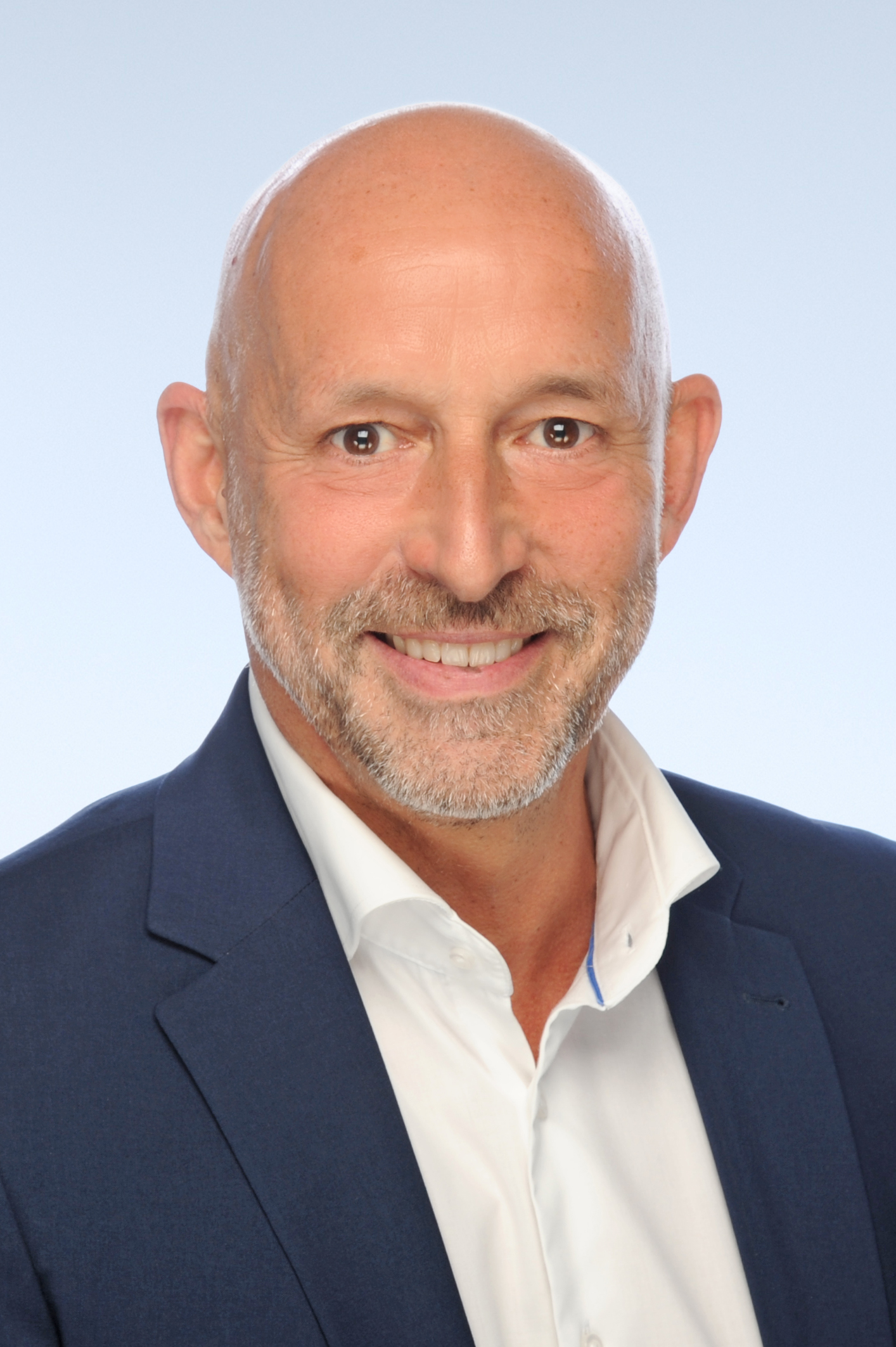 Portraitfoto eines Mannes mit Glatze und kurzem Bart in einem weißen Hemd und dunkelblauer Anzugjacke, der freundlich in die Kamera lächelt.