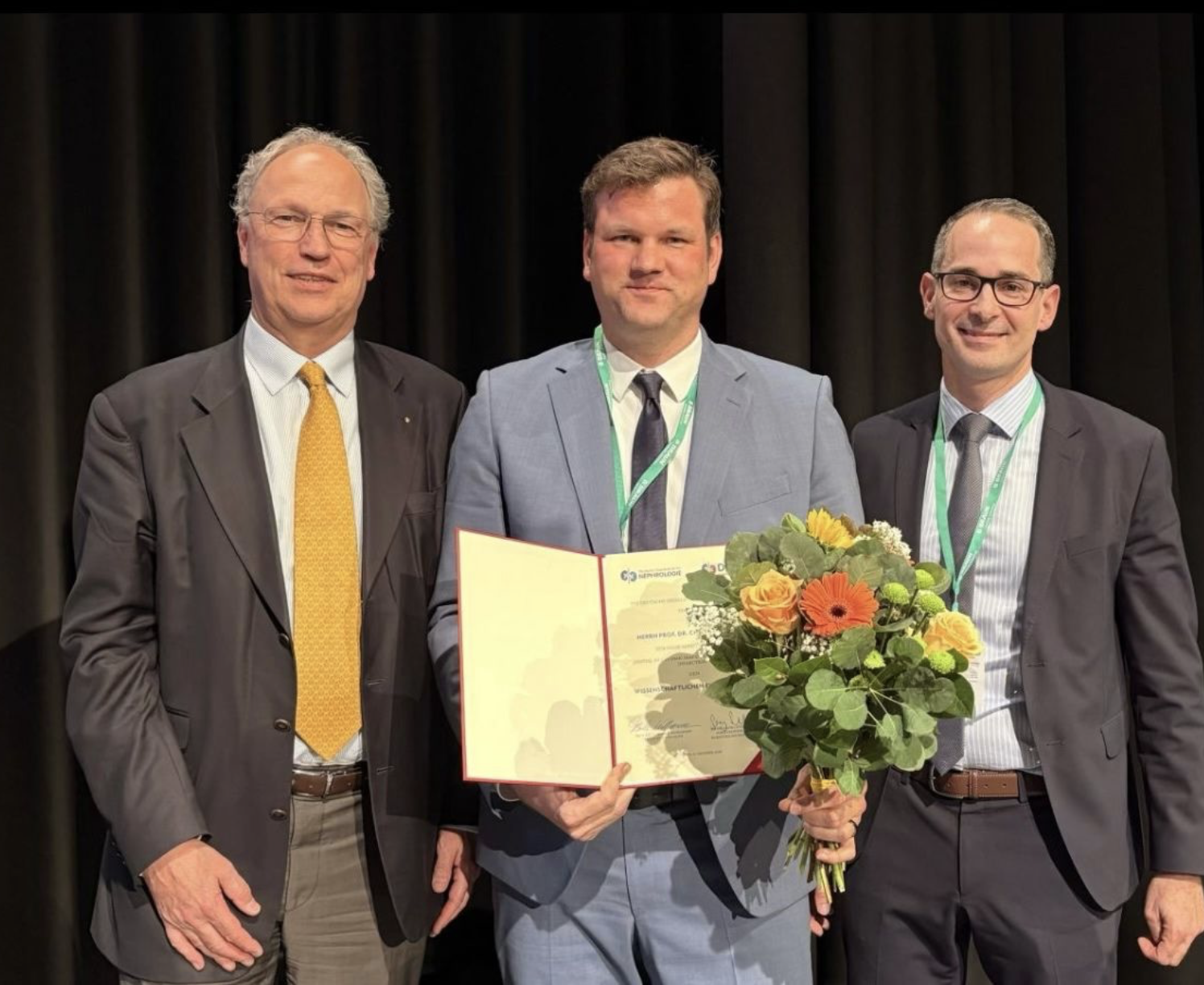 Gruppenfoto dreier Männer mittleren Alters in Anzügen. Der Mann in der Mitte hält eine Urkunde und einen Blumenstrauß.