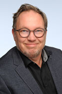 Dipl.-Pflegew. (FH) Mario Hoben: Ein lächelnder Mann mit Brille, dunklem Blazer über einem schwarzen Hemd. Der Hintergrund ist hellblau.