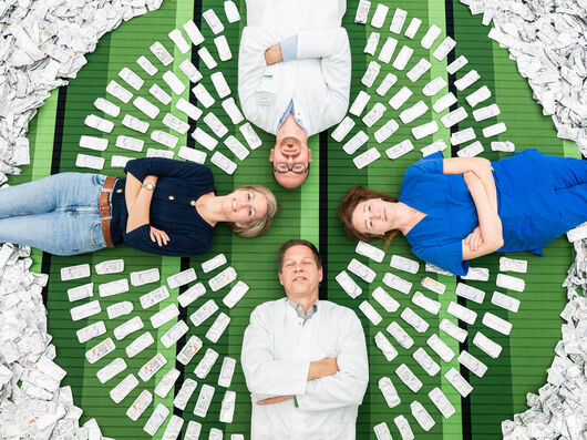 Vier Personen liegen in Kreuzform auf einer grünen Fläche, umgeben von zahlreichen aluminiumhaltigen Nahtverpackungen, die kreisförmig um sie angeordnet sind.