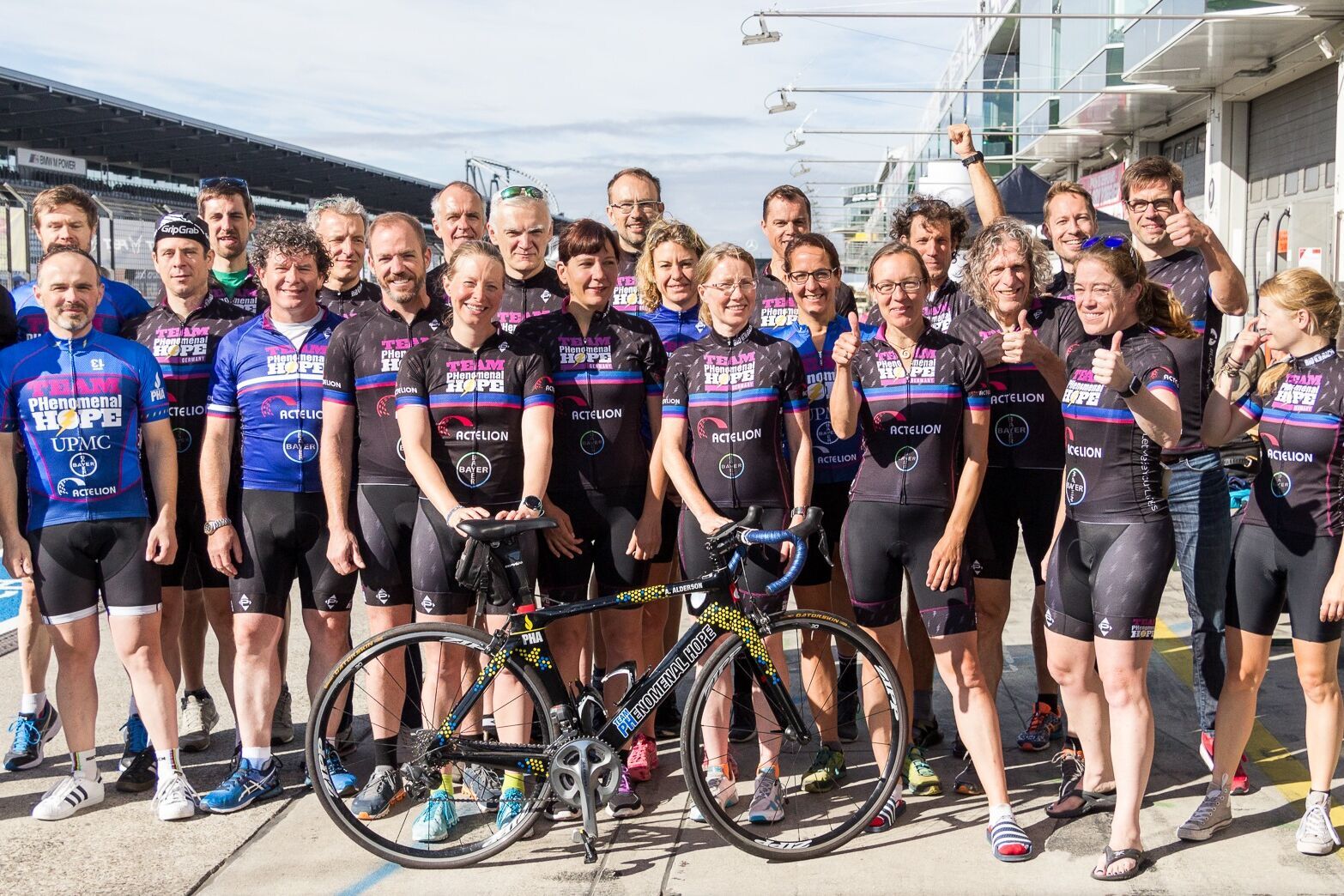 Das Bild zeigt eine Gruppe von Radfahrern, die zusammen posieren und passende Radtrikots tragen. Im Vordergrund befindet sich ein Fahrrad.