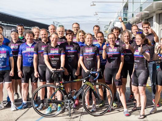 Das Bild zeigt eine Gruppe von Radfahrern, die zusammen posieren und passende Radtrikots tragen. Im Vordergrund befindet sich ein Fahrrad.