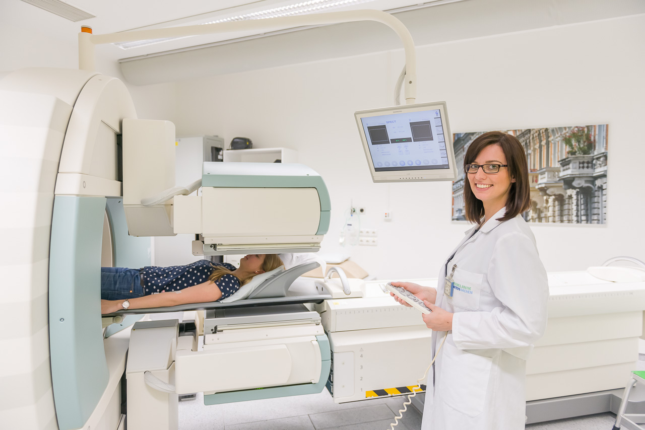 Medizinische Umgebung, in der ein Patient auf einem Bett liegt und teilweise in einem großen medizinischen Bildgebungsgerät ist. Ein medizinischer Fachmann im weißen Kittel steht in der Nähe, schaut auf einen Monitor und hält ein Tablet oder Klemmbrett. Der Monitor zeigt einige Daten oder Bilder, die mit dem Verfahren zusammenhängen.