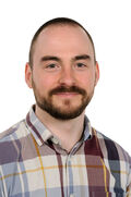 Portraitfoto eines Mannes mit kurzen braunen Haaren und Bart in einem karierten Hemd, der freundlich in die Kamera blickt.