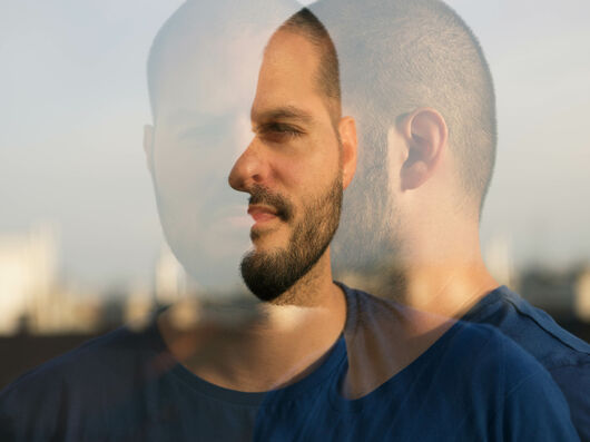 Mann steht vor Gebäude mit Doppelbelichtung, also sieht man ihn einmal von vorne und einmal von der Seite. Er trägt einen Buzzcut und hat einen Fünftagebart.