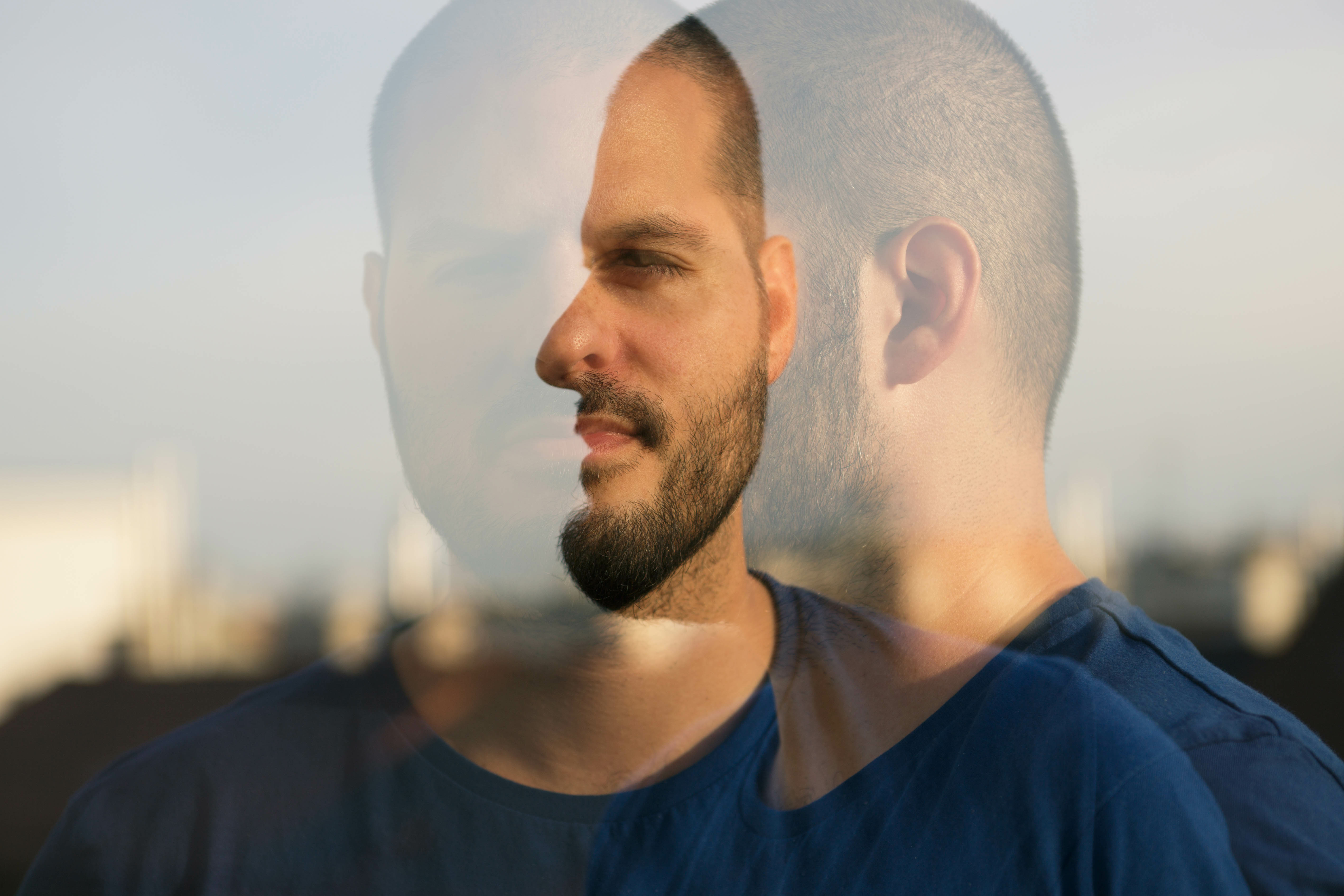 Mann steht vor Gebäude mit Doppelbelichtung, also sieht man ihn einmal von vorne und einmal von der Seite. Er trägt einen Buzzcut und hat einen Fünftagebart.