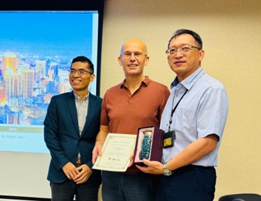 [Translate to Englisch:] Auf dem Bild sind drei Männer zu erkennen. Der Mann links trägt einen Anzug und schaut nach links. Die anderen zwei Männer tragen Hemden und schauen zur Kamera. Der Mann in der Mitte hält eine Urkunde. 