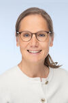 Portraitfoto einer Frau mit hellbraunen Haaren und runder Brille in einem hellgrauen Pullover, die freundlich in die Kamera lächelt.