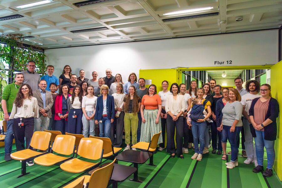 Auf dem Foto erkannt man das Team des Zentrums für Humangenetik und Genommedizin. Der Boden ist grün, und es gibt mehrere gelbe Stühle im Raum. Im Hintergrund sieht man eine weiße Wand mit einem Schild über einer offenen Tür. Auf dem Schild steht „Flur 12“. Auf der linken Seite des Bildes sind einige Pflanzen zu sehen.
