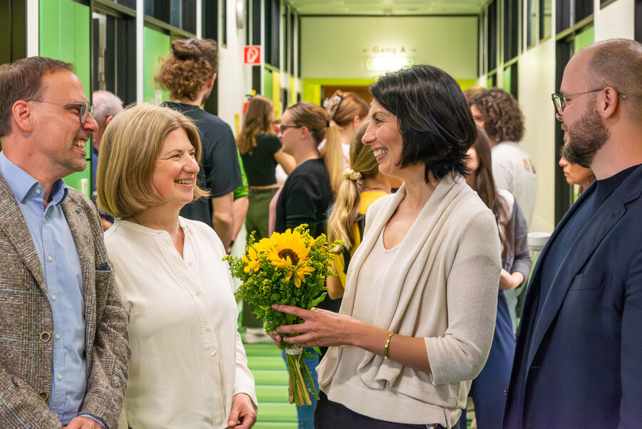 Das Bild zeigt eine Gruppe von Menschen, die in einem Flur stehen. Im Vordergrund sind vier Personen zu sehen. Eine dieser Personen hält einen Blumenstrauß mit gelben Blumen, darunter auch Sonnenblumen. Im Hintergrund stehen weitere Menschen.
