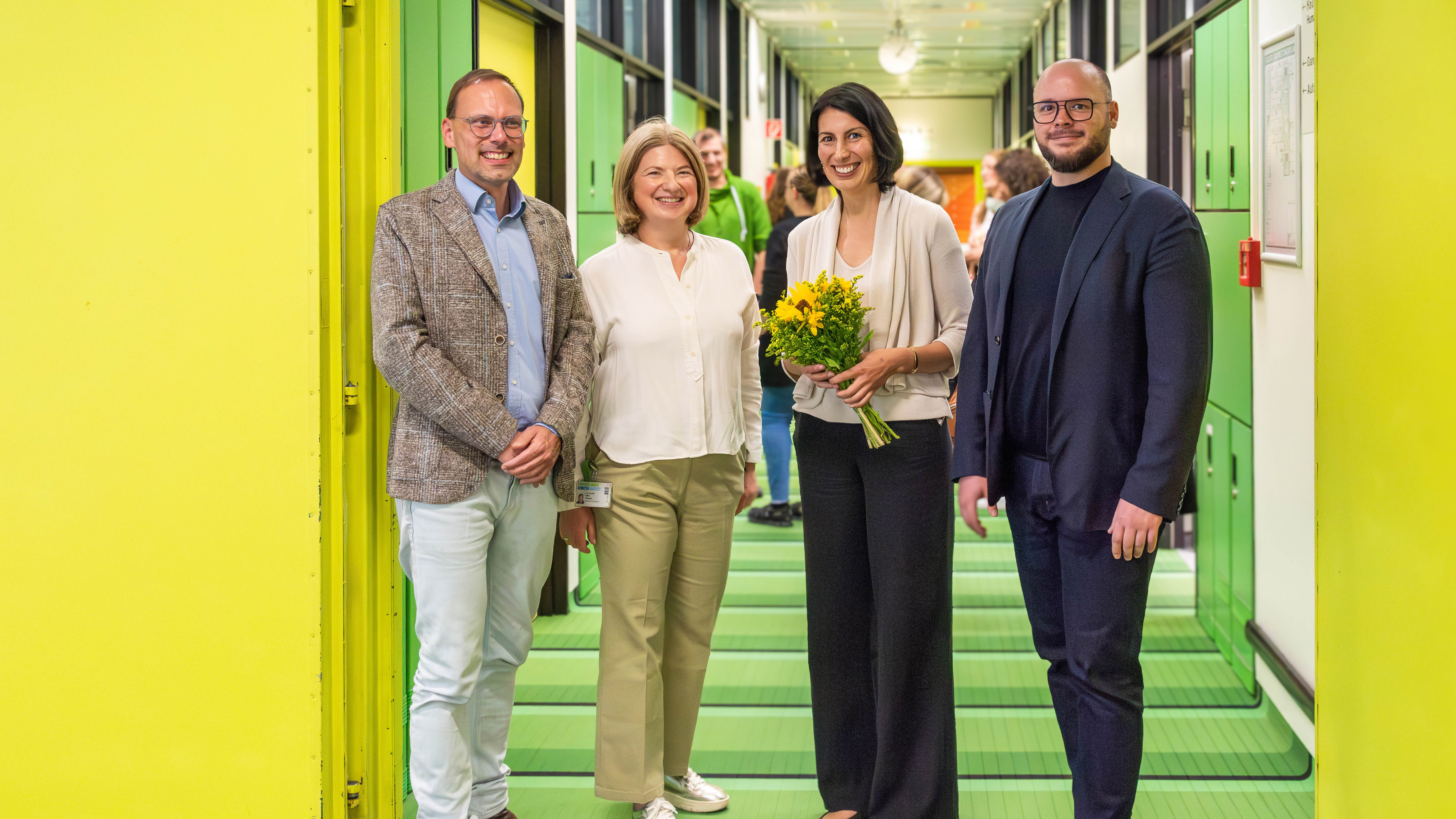 Vier Personen stehen in einem Flur mit grünen Wänden und grünem Boden. Eine Person hält einen Strauß gelber Blumen.