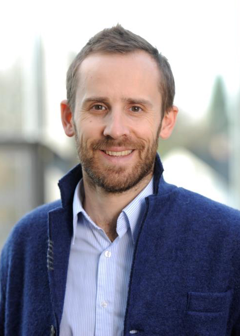 Portraitfoto eines Mannes mit kurzen braunen Haaren in einem hellblauen Hemd und dunkelblauem Jackett, der freundlich in die Kamera lächelt.