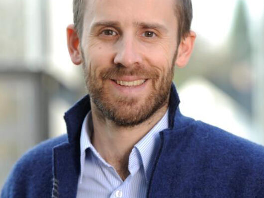 Portraitfoto eines Mannes mit kurzen braunen Haaren in einem hellblauen Hemd und dunkelblauem Jackett, der freundlich in die Kamera lächelt.