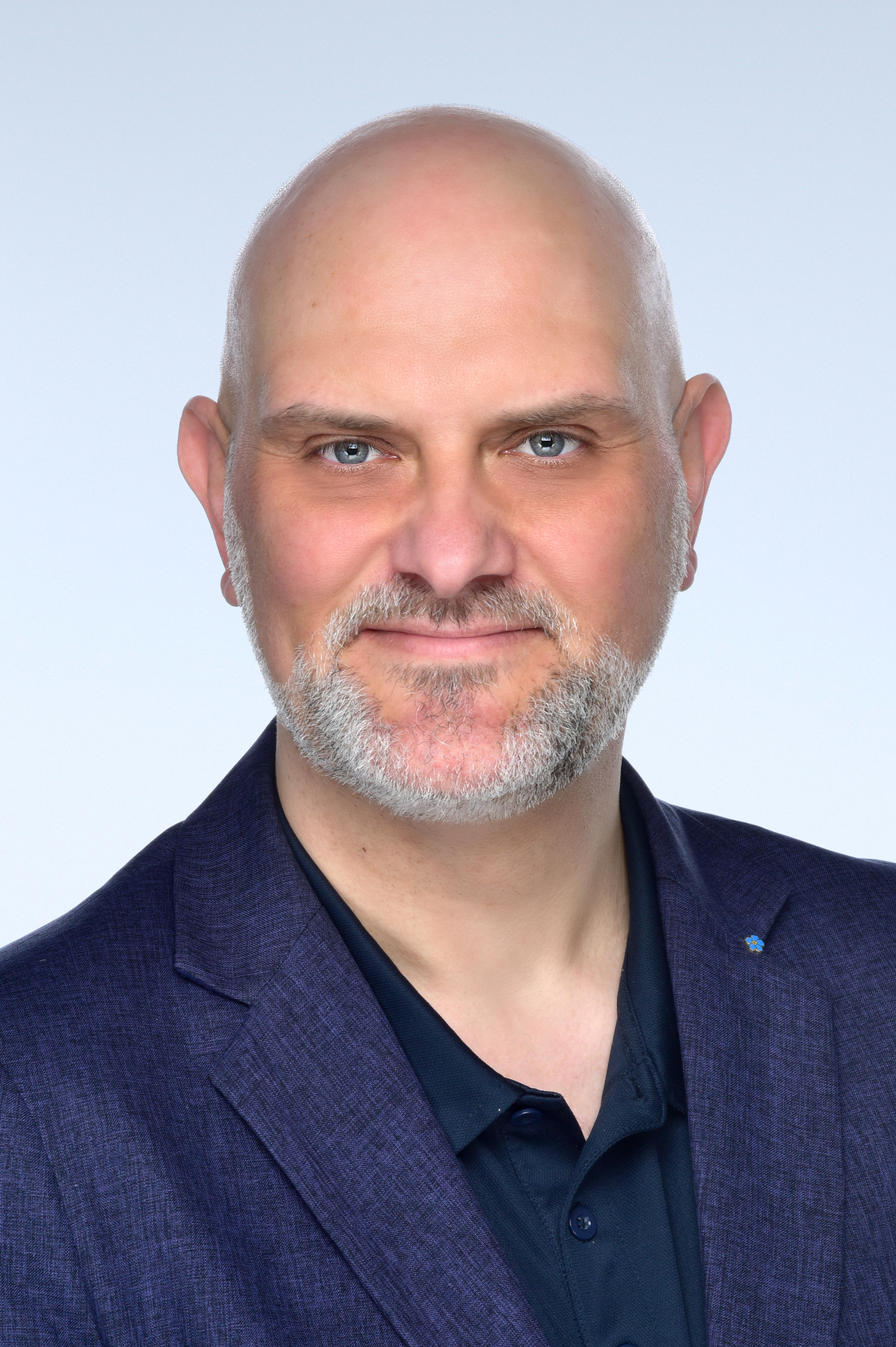 Ein mittelalter, weißer Mann mit grauem Bart und blauen Augen. Er trägt ein blaues Sakko.