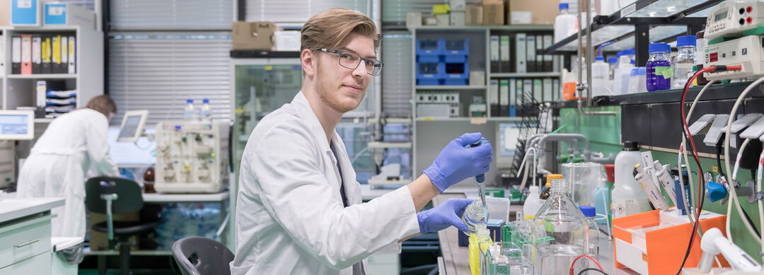 Bild eines jungen Mannes mit kurzen braunen Haaren und Brille in weißer Laborkleidung, der an einem Arbeitsplatz sitzt und pipettiert. Auf der Tischplatte stehen diverse Flüssigkeiten in Flaschen.