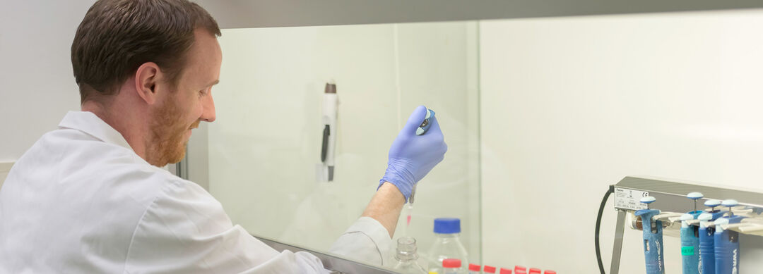 Bild eines Mannes mit kurzen braunen Haaren in weißem Laborkittel beim Pipettieren unter einem Abzug. 