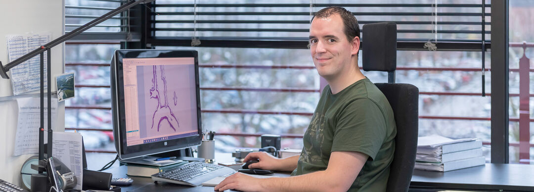 Bild eines Mannes mit kurzen braunen Haaren in einem grünen T-Shirt, der am Schreibtisch sitzt und auf ein Bild einer Probe schaut.