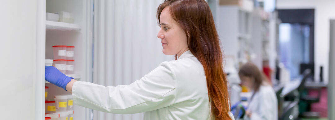 Bild einer Frau mit langen braunen Haare in einem weißen Laborkittel vor einem geöffneten Kühlschrank, in den sie eine Probe legt.