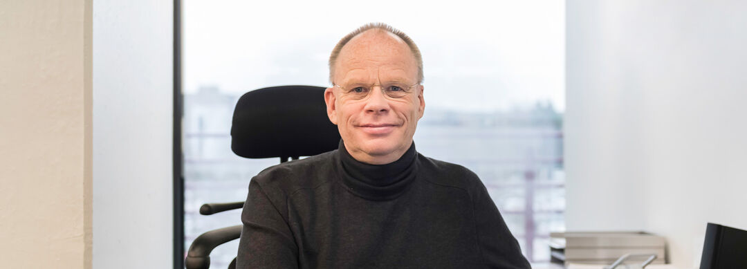 Bild eines Mannes mit Glatze und randloser Brille in einem schwarzen Rollkragenpullover am Schreibtisch sitzend.