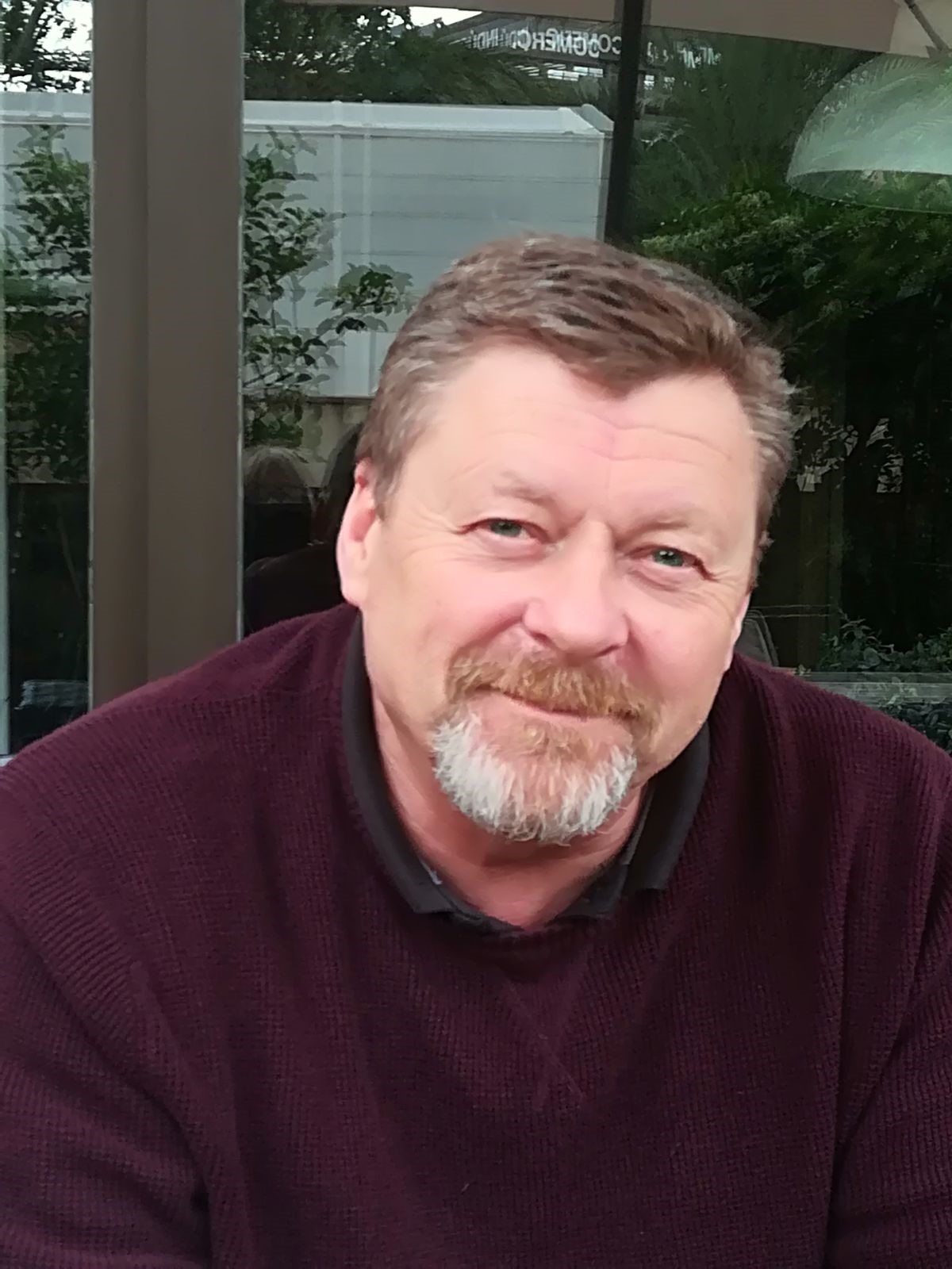 Ein älterer Herr mit angegrauten Haaren und Bart. Er trägt einen purpurnen Pullover.