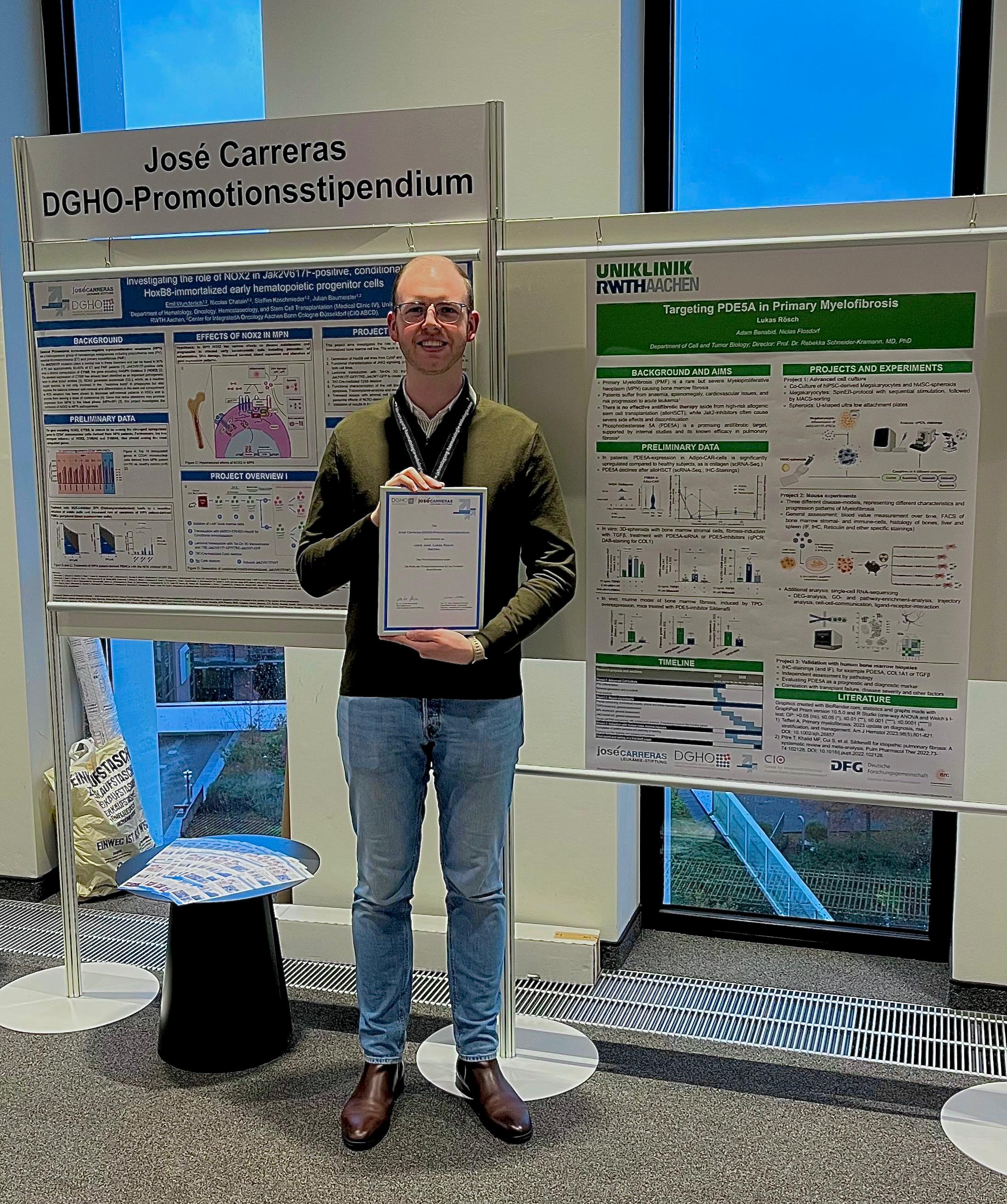[Translate to Englisch:] Mann hält eine Urkunde und steht vor zwei wissenschaftlichen Postern.