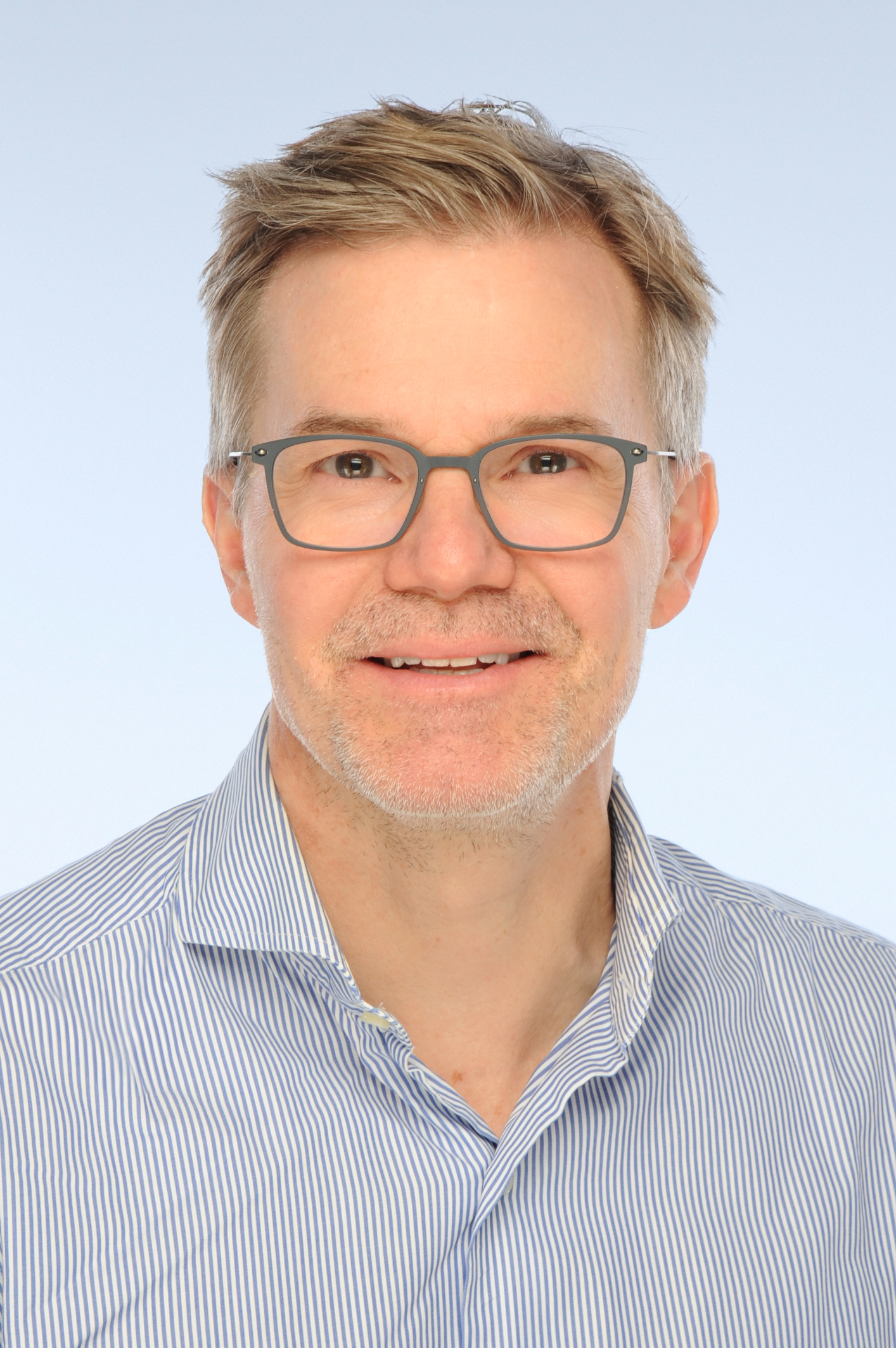 Porträtfoto eines Mannes mit kurzen Haaren und einer Brille. Er trägt ein gestreiftes Hemd, hat einen Bart und lächelt freundlich in die Kamera. 