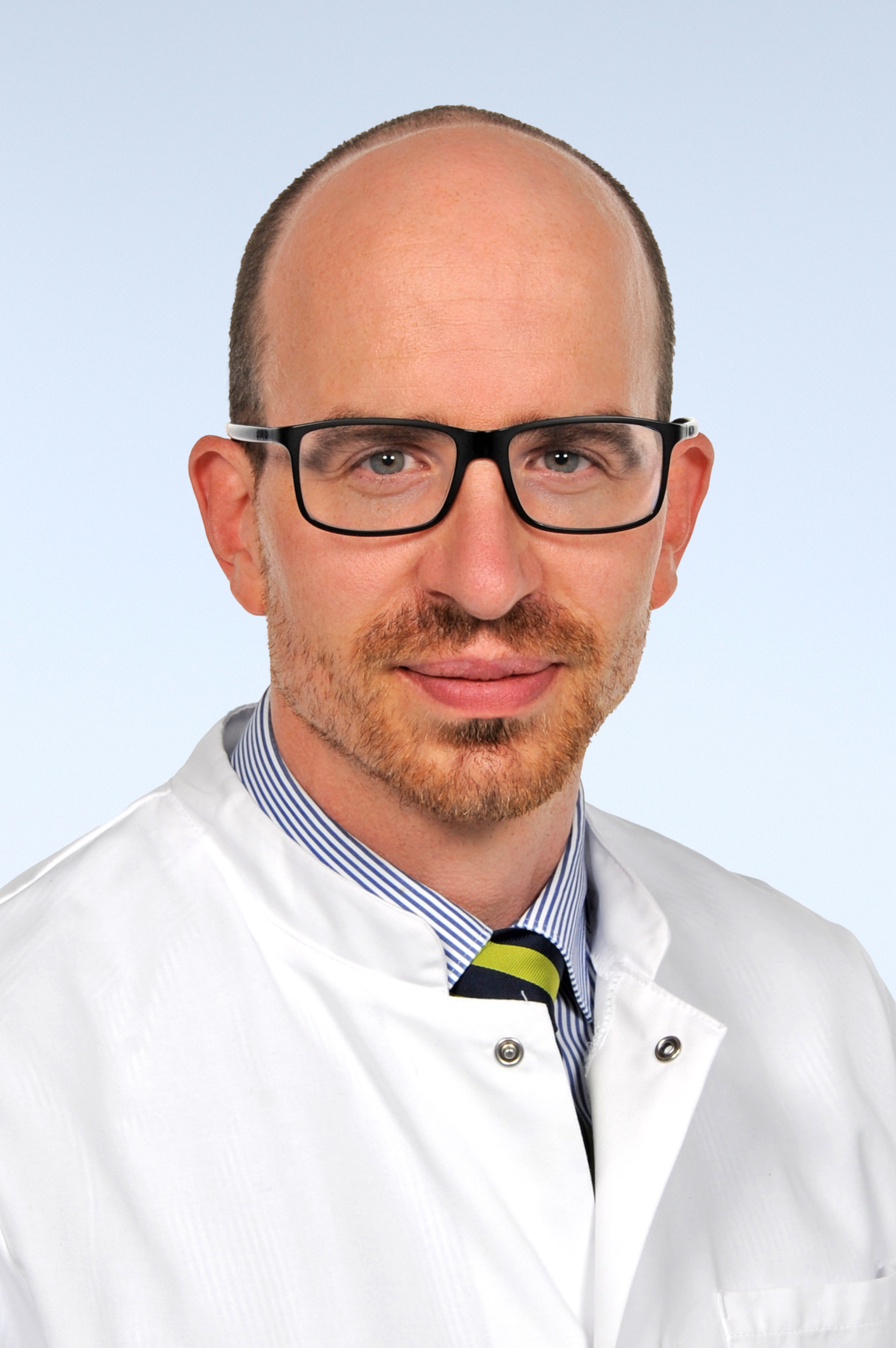 Porträt eines Mannes, etwa Mitte 40, mit kurzen braunen Haaren, trägt eine schwarze Brille. Er trägt einen weißen Arztkittel über einem hellblauen Hemd und lächelt freundlich.