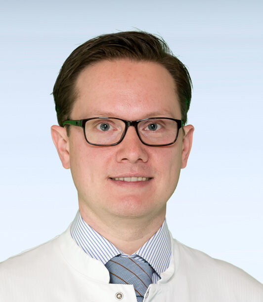 Portraitfoto eines Mannes mit kurzen dunkelbraunen Haaren und eckiger Brille, der in die Kamera lacht.