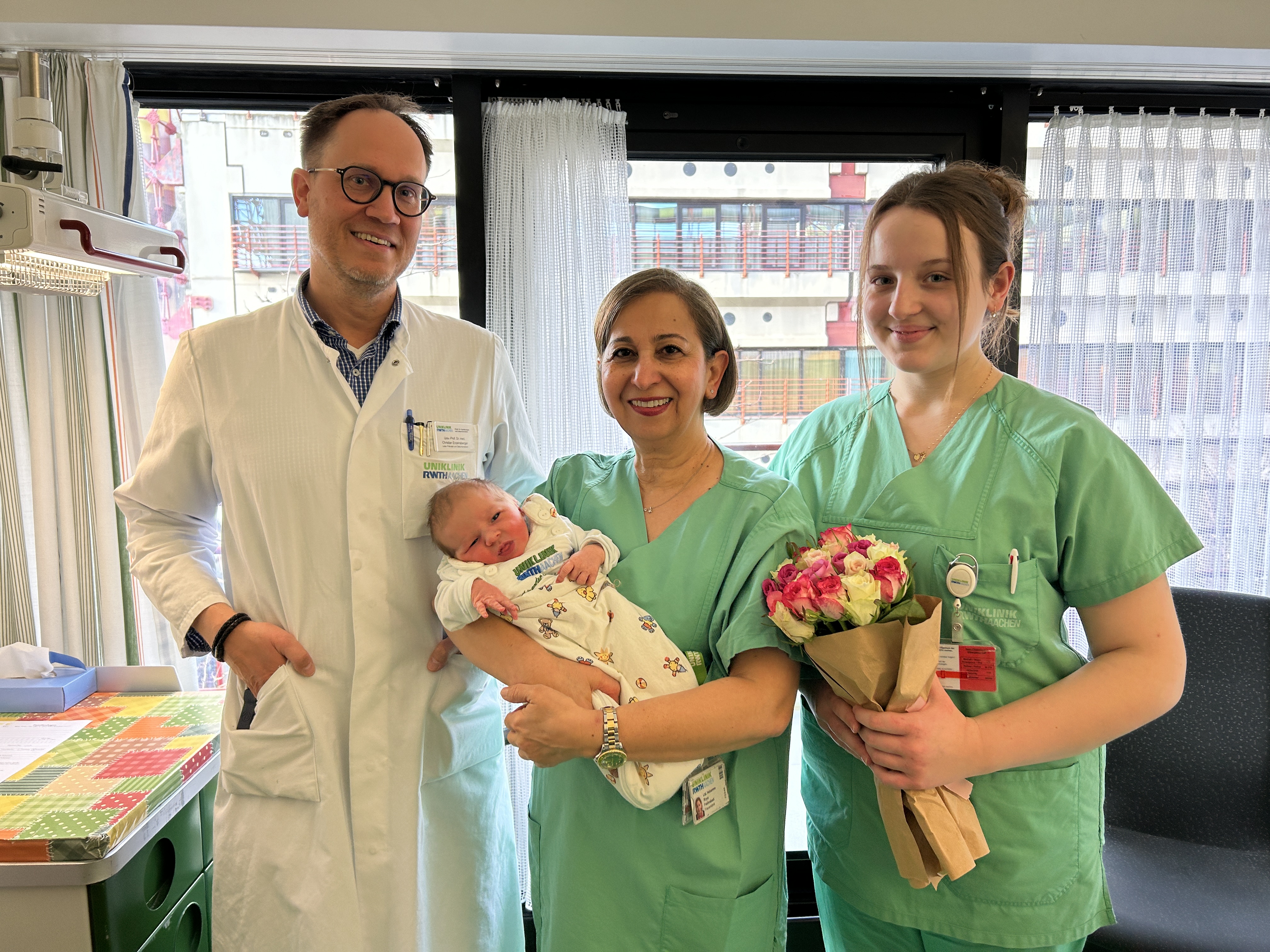 Ein Mann mit Brille in einem weißen Arztkittel steht neben zwei Frauen in grünem Kasack. Die Dame in der Mitte hat kürzere dunkle Haare und hält ein Baby auf dem Arm. Die Dame rechts hat längere dunklere Haare und hält einen Blumenstrauß.