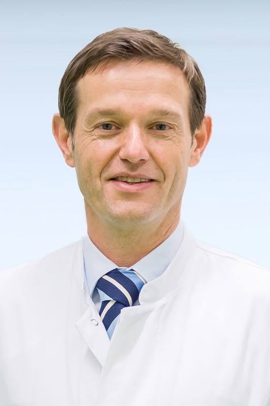 Porträtfoto eines Mannes mit braunen Haaren. Er trägt einen weißen Arztkittel und eine blau-gestreifte Krawatte. Er lächelt freundlich in die Kamera. 