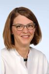Porträtfoto einer Frau mit braunen Haaren und einer Brille in einem weißen Arztkittel. Sie lächelt freundlich in die Kamera. 