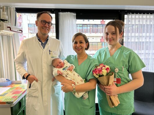 Ein Mann mit Brille in einem weißen Arztkittel steht neben zwei Frauen in grünem Kasack. Die Dame in der Mitte hat kürzere dunkle Haare und hält ein Baby auf dem Arm. Die Dame rechts hat längere dunklere Haare und hält einen Blumenstrauß.