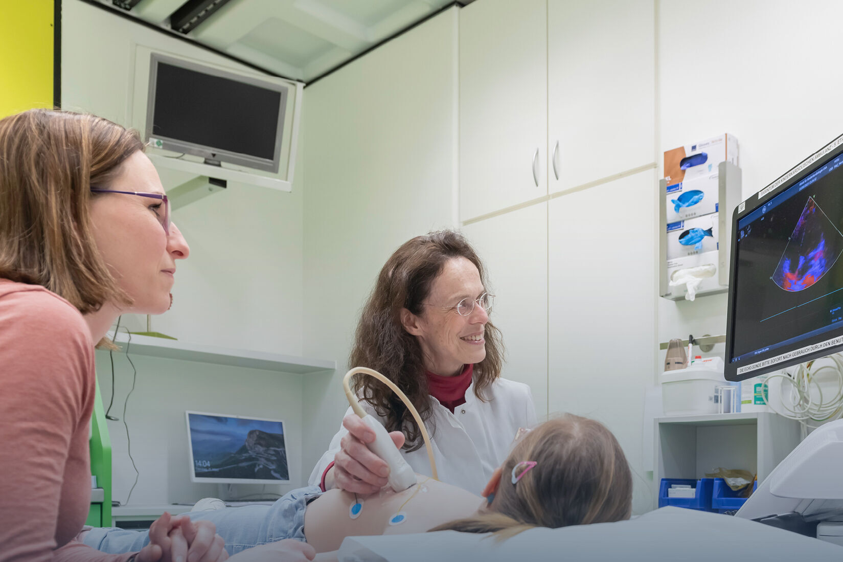 Das Bild zeigt eine medizinische Umgebung, in der eine Frau in einem weißen Arztkittel einen kleinen Patienten mit dem Ultraschallgerät untersucht.