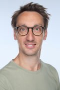 Porträtfoto eines Mannes mit braunen Haaren und einer Brille mit runden Brillengläsern.