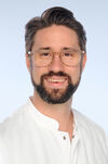 Porträtfoto eines Mannes mit dunklen Haaren und Bart. Er lächelt und trägt einen weißen Arztkittel.