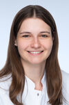 Portraitfoto einer Frau mit langen braunen Haaren, die freundlich in die Kamera lächelt und einen Arztkittel trägt.