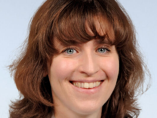 Porträt einer Frau mit schulterlangen rotbraunen Haaren. Sie lächelt und trägt einen weißen Arztkittel.