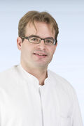 Porträtfoto eines Mannes mit kürzeren, dunkelblonden Haaren und einer Brille mit dunklem Brillengestell. Er trägt einen weißen Arztkittel und lächelt freundlich in die Kamera. 
