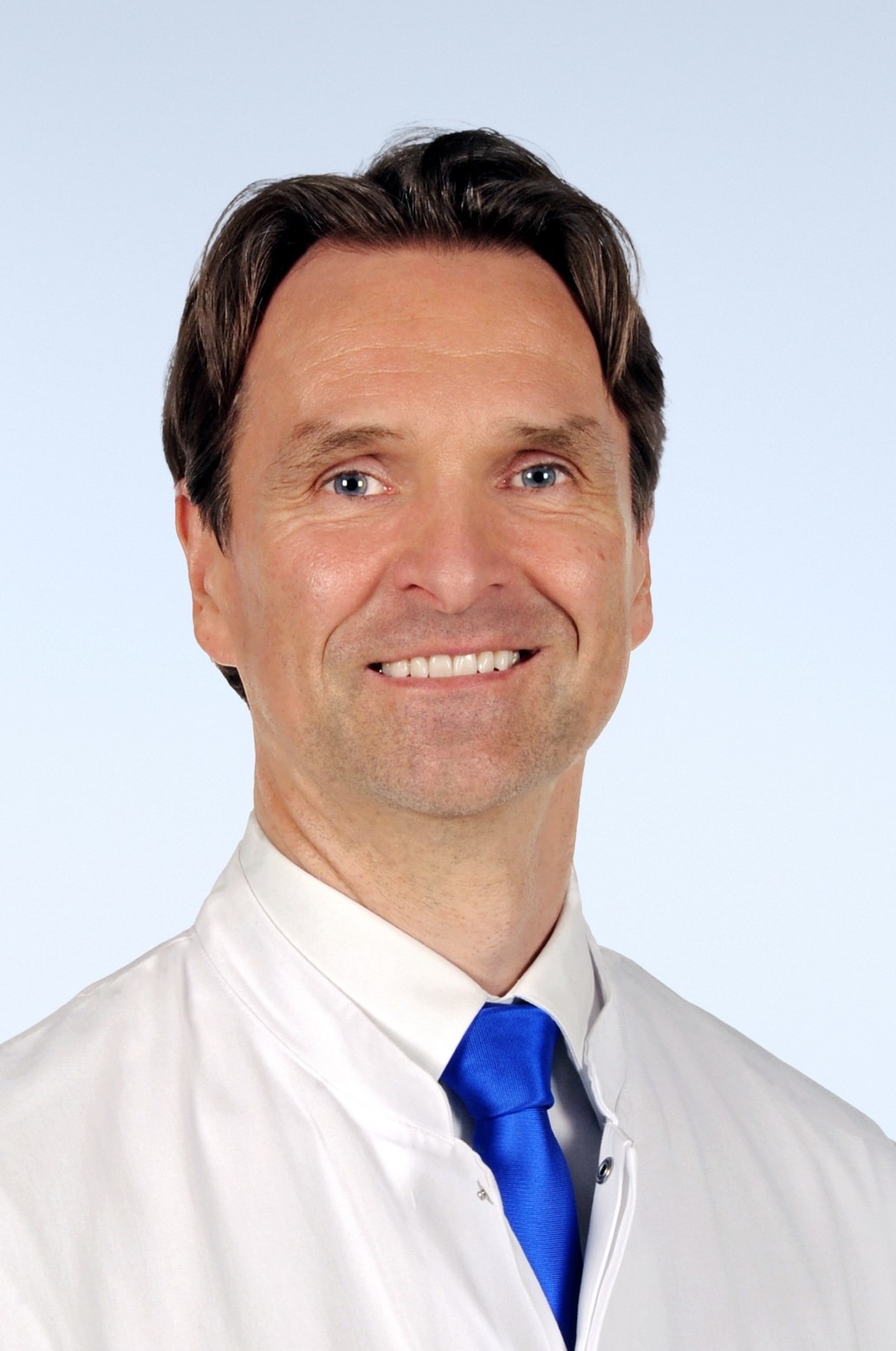 Portraitfoto von Mann mit kurzen braunen Haaren, weißem Arztkittel, weißem Hemd und blauer Krawatte, grinst in die Kamera